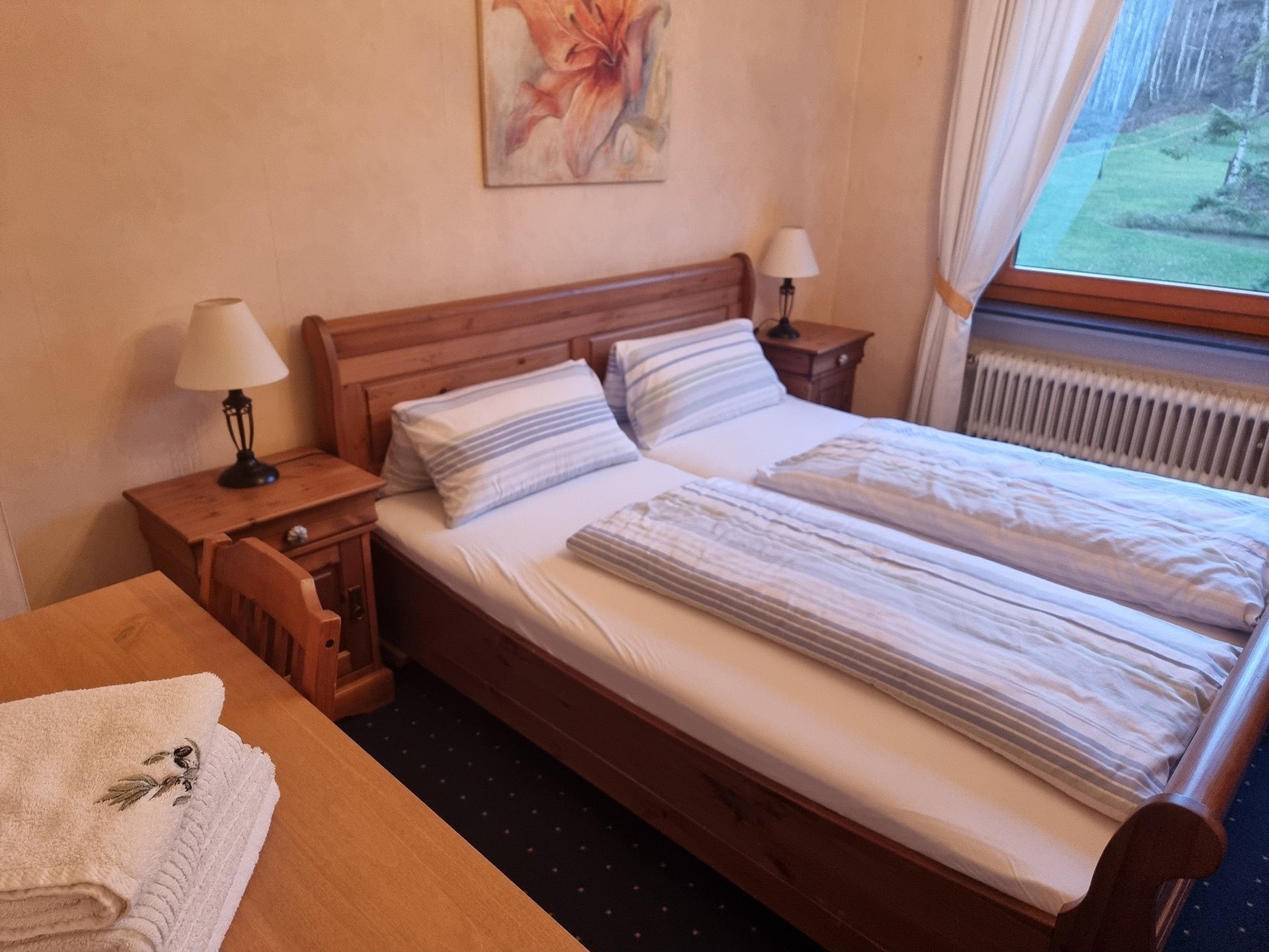 Zimmer ‚Prato‘ in der Pension Rehwinkel in Nalbach, mit rustikalem Holzbett, gemütlicher Einrichtung, einem Blumenbild und großem Fenster mit Ausblick ins Grüne.